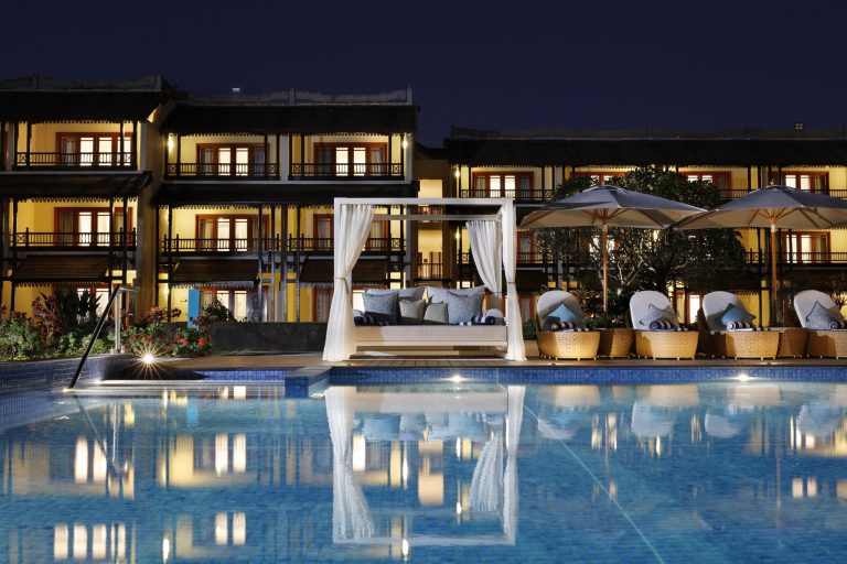 Poolbereich mit blau gekacheltem Wasser, Liegestühlen mit blauen und weißen Kissen und Baldachin-Bett vor dreistöckigem.