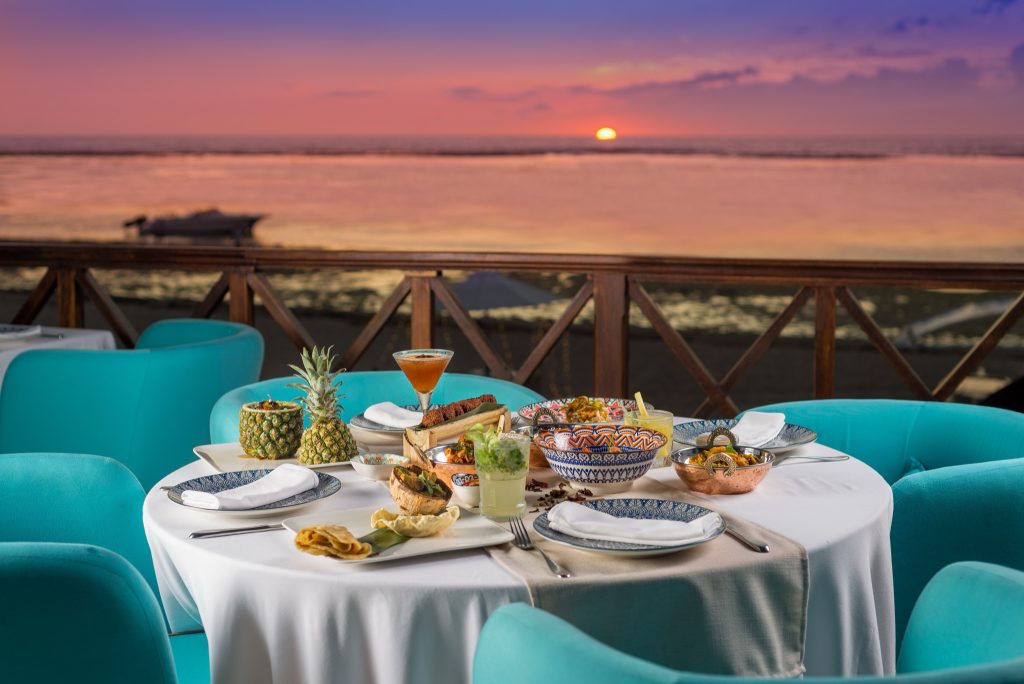 Tisch im Restaurant Jacaranda mit weißem Tischtuch, türkisfarbenen Stühlen und Meerblick bei Sonnenuntergang