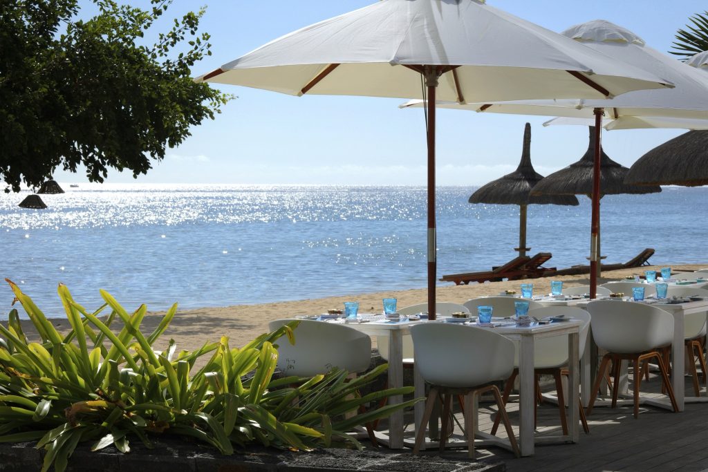 Strandterrasse des Restaurants Tamassa mit weißen Tischen und Stühlen unter Sonnenschirmen und Blick auf das Meer