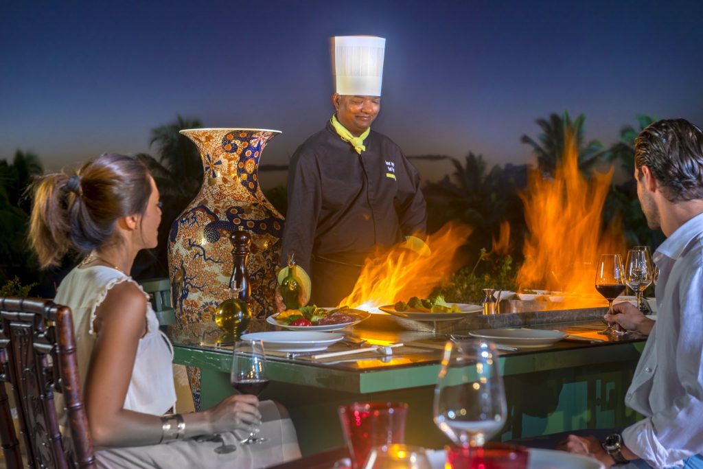 Gehobenes Restaurant mit Teppanyaki-Koch am Grill, zwei Gästen, Tisch mit Keramikgefäß und Gläsern bei Abenddämmerung