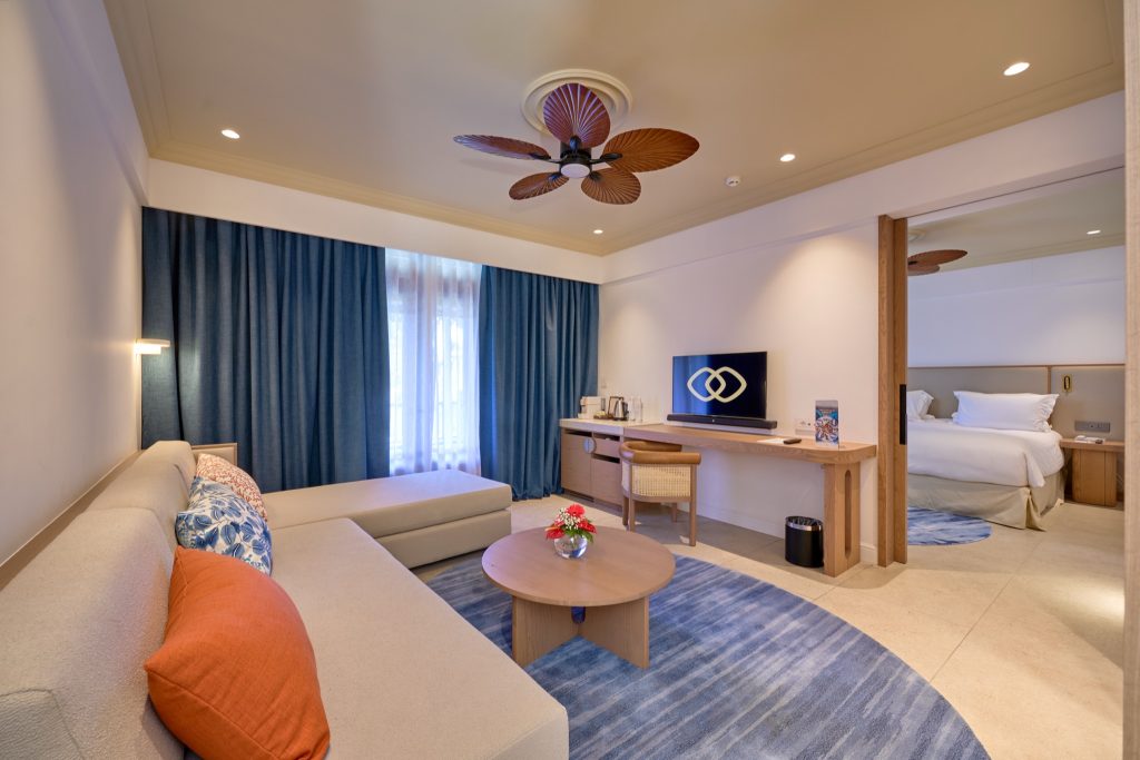Modernes Family Suite Zimmer mit beige Sofa, bunten Kissen, blauem Teppich und angrenzendem Schlafzimmer