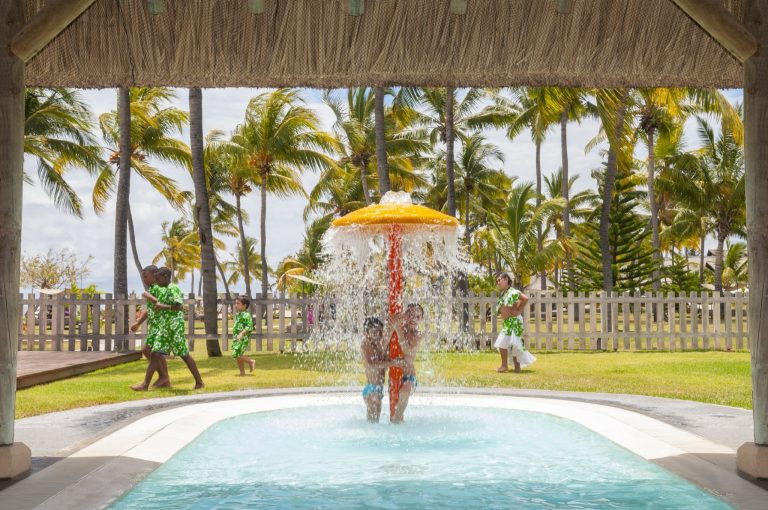 Spielbereich mit orangefarbenem Regenschirm als Wasserquelle und kleinem Pool umgeben von Palmen auf grünem Rasen