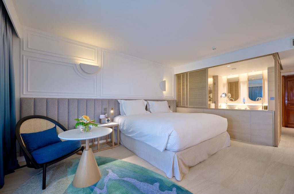 Modernes Hotelzimmer mit Doppelbett, Nachttisch, Sitzgelegenheit und Holzlamellen-Trennwand zum Bad