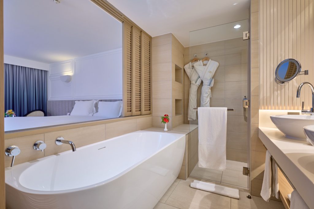 Hotelzimmer mit freistehender Badewanne, Glasdusche, Holzverkleidung und Blick auf das Bett mit blauen Vorhängen