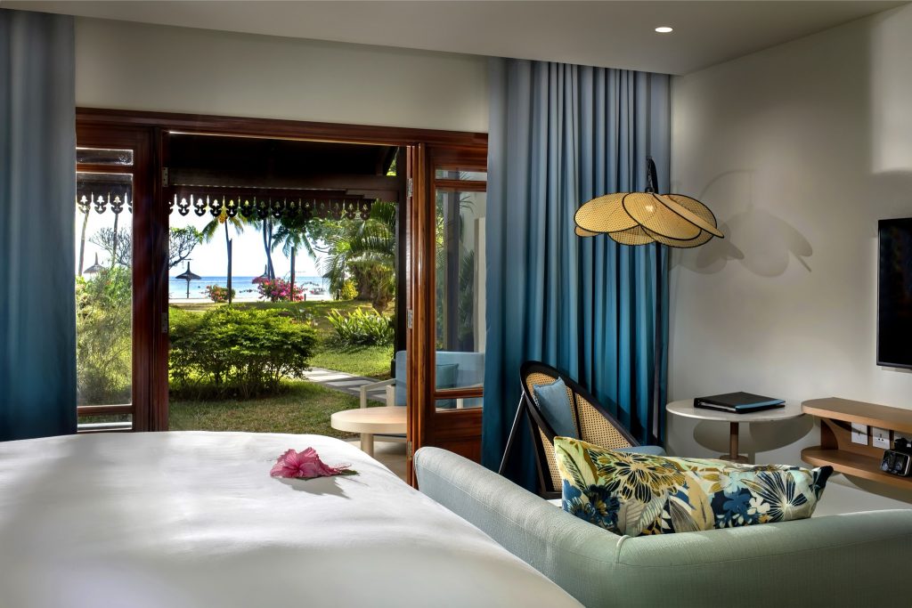Hotelzimmer Magnifique Room mit großem Bett, pinkfarbener Blüte, Sofa, Rattanstuhl und Meerblick durch geöffnete Glastür