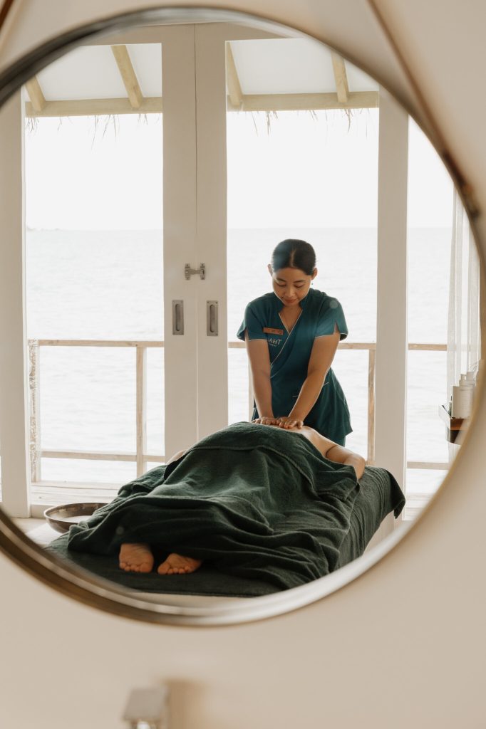 Massage im Spa mit blauer Uniform, dunkelgrüner Massagebank, rundem Spiegel und Meerblick durch