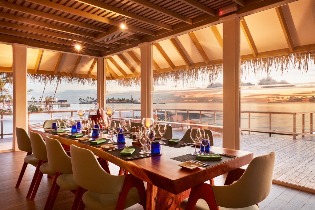 Bar Wine Cellar im Sun Siyam Iru Veli mit Holztisch, cremefarbenen Stühlen und Blick auf den Sonnenuntergang über dem Meer