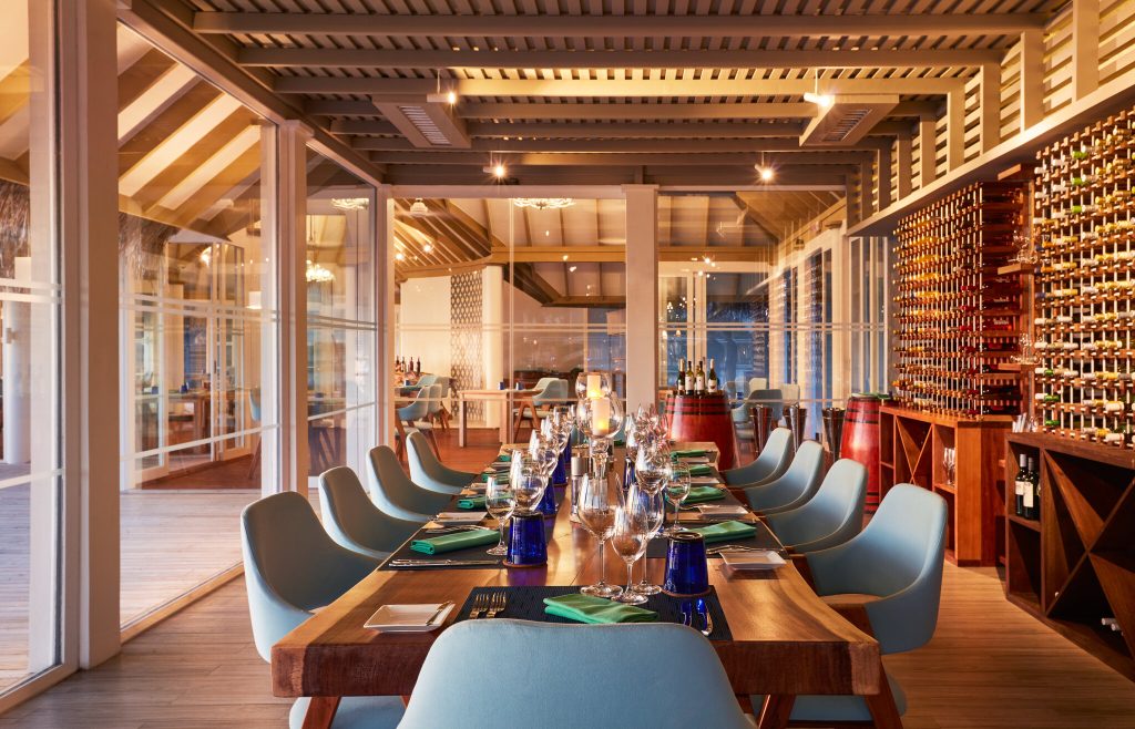 Bar Wine Cellar mit langem Holztisch, blauen Gläsern, hellblauen Stühlen und Weinregalen aus Holz