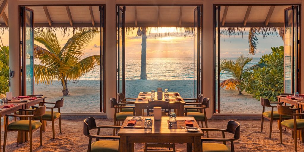 Offenes Strandrestaurant Aqua Orange mit Holztischen und grünen Kissen, Blick auf Meer und Palmen bei Sonnenuntergang