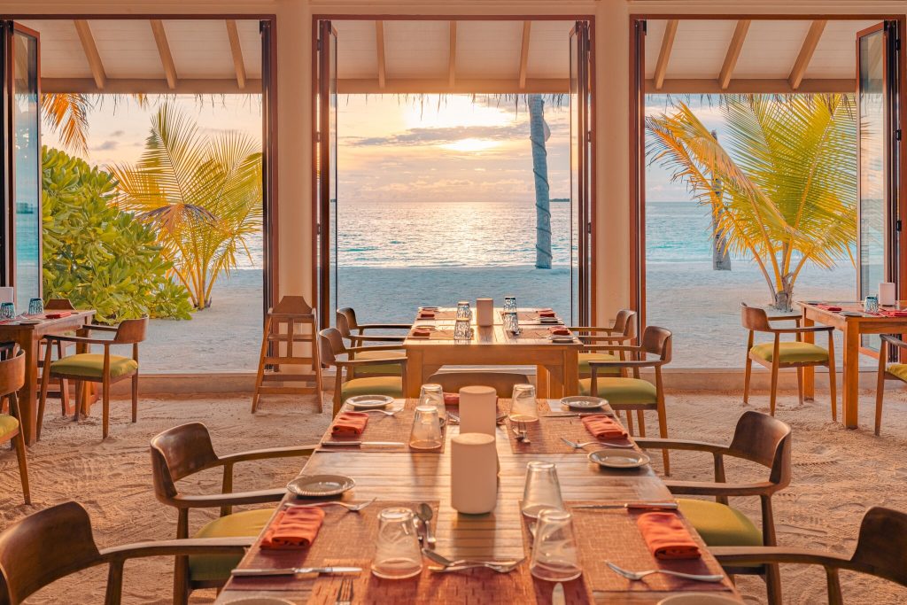 Restaurant aqua orange mit Holztischen, grünen Stühlen und Blick auf Strand und Meer bei Sonnenuntergang
