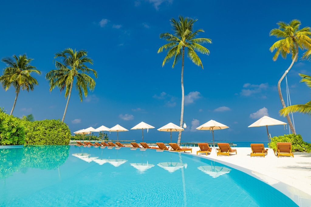 Infinity-Pool mit Holzliegen und cremefarbenen Sonnenschirmen auf weißem Sand, Palmen und blauer Himmel im Hintergrund