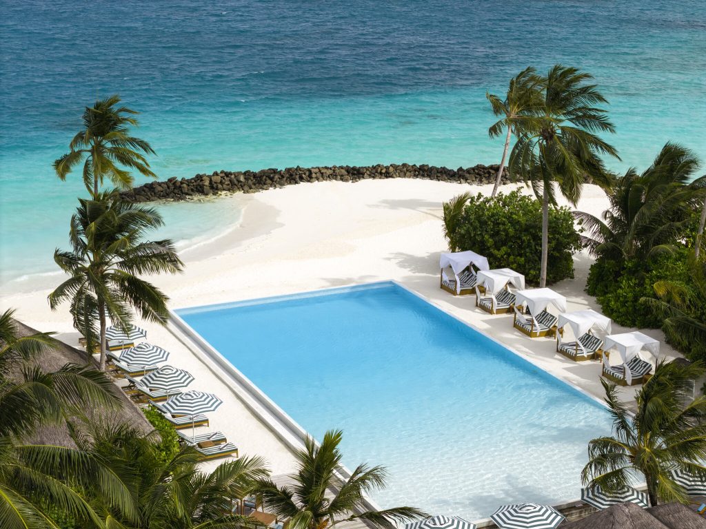 Großer rechteckiger Pool mit weißen Liegen und gestreiften Kissen, umgeben von Palmen und weißem Sandstrand am Meer