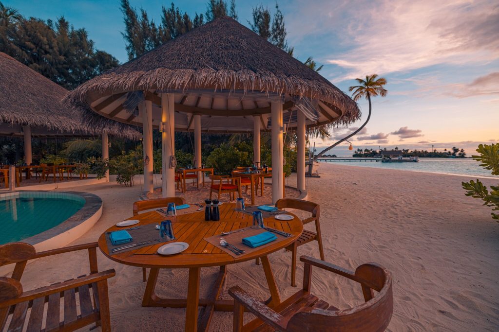 Offene Hotelterrasse mit rundem Holztisch und blauen Servietten, Strohdach-Pavillons und Meer im Hintergrund