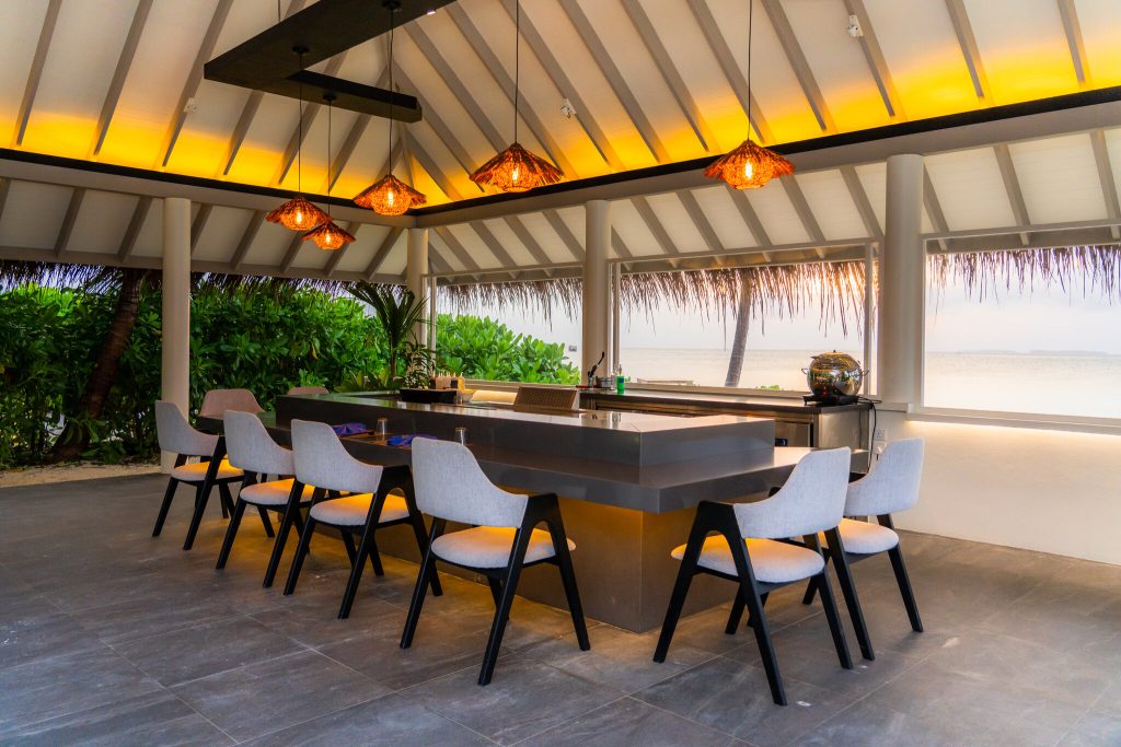 Offene Hotelbar mit modernem Tresen, Holzstühlen mit hellen Sitzflächen und Blick auf tropische Vegetation und Meer