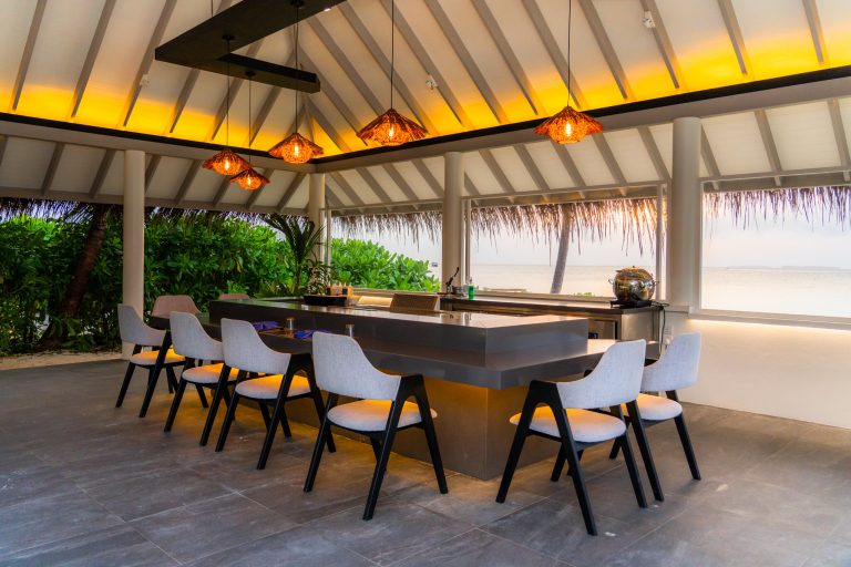 Offene Hotelbar mit modernem Tresen, Holzstühlen mit hellen Sitzflächen und Blick auf tropische Vegetation und Meer