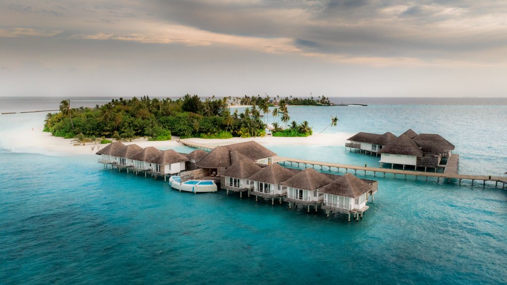 Bungalows auf Stelzen mit Strohdächern über türkisblauem Wasser und grüner Insel im Hintergrund