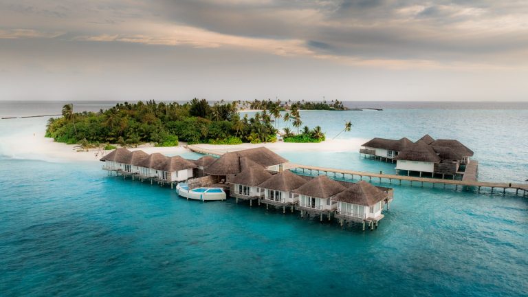 Bungalows auf Stelzen mit Strohdächern über türkisblauem Wasser und grüner Insel im Hintergrund