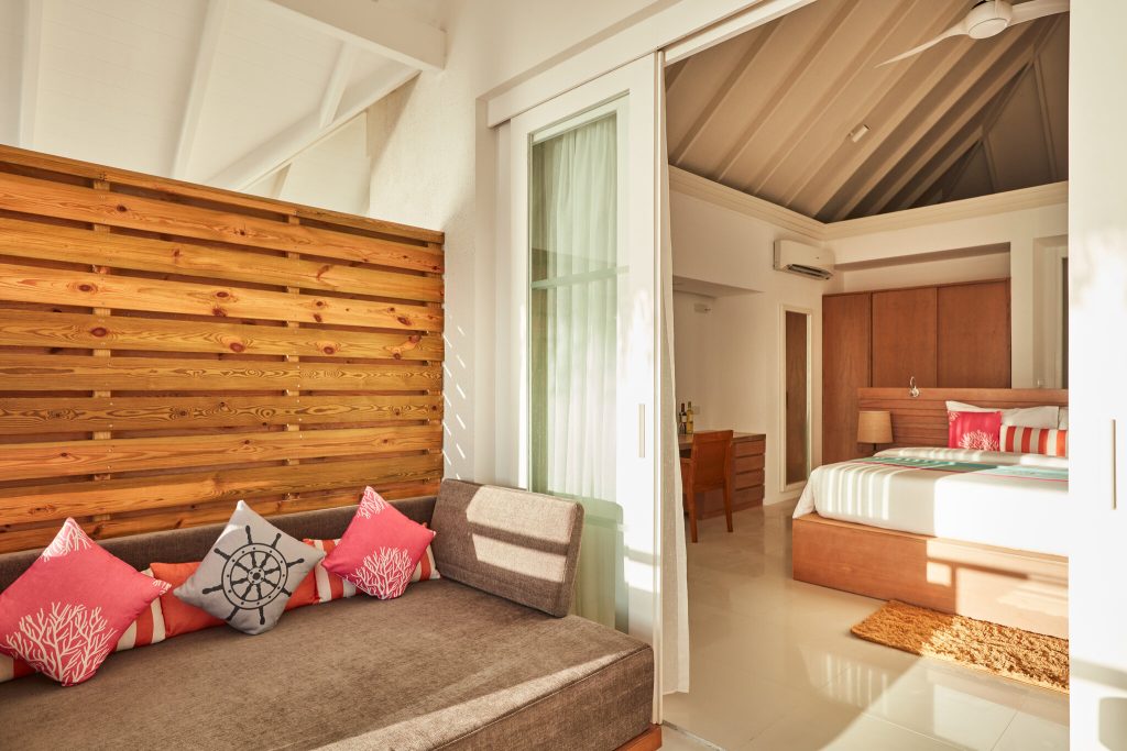 Hotelzimmer Beach Residence mit Holzvertäfelung, roter Couch und Schlafzimmer hinter Glastür im Sun Siyam Iru Veli