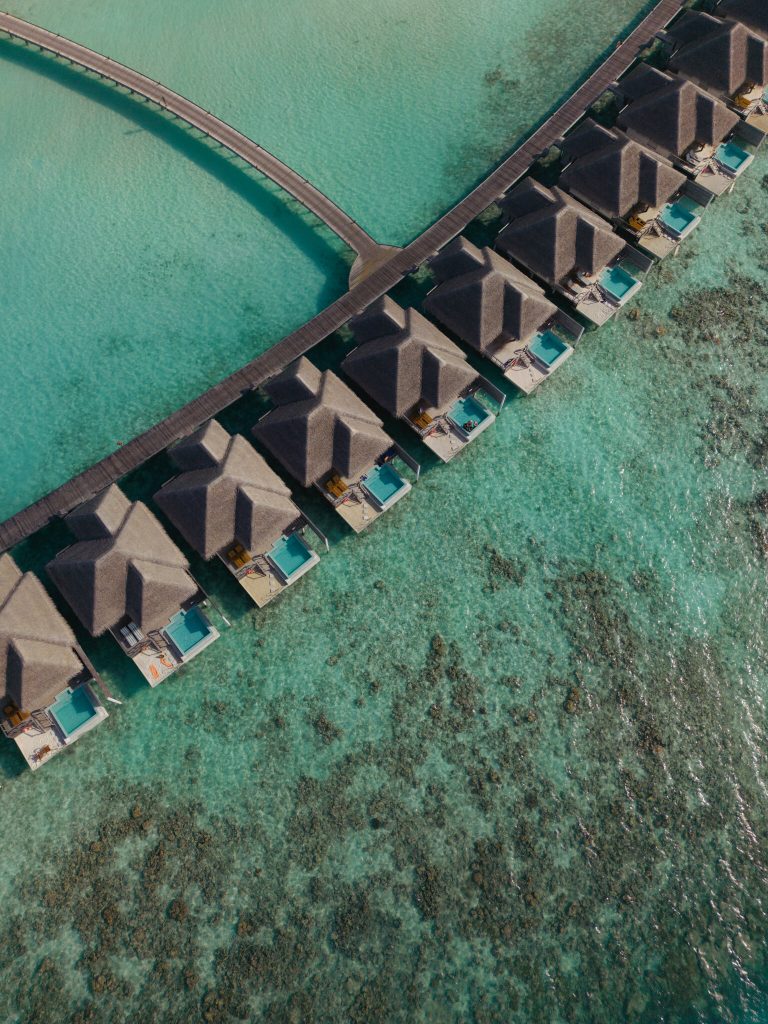 Reihe von Dolphin Ocean Villas mit Strohdächern auf Stelzen über türkisfarbenem Wasser, verbunden durch hölzernen Steg