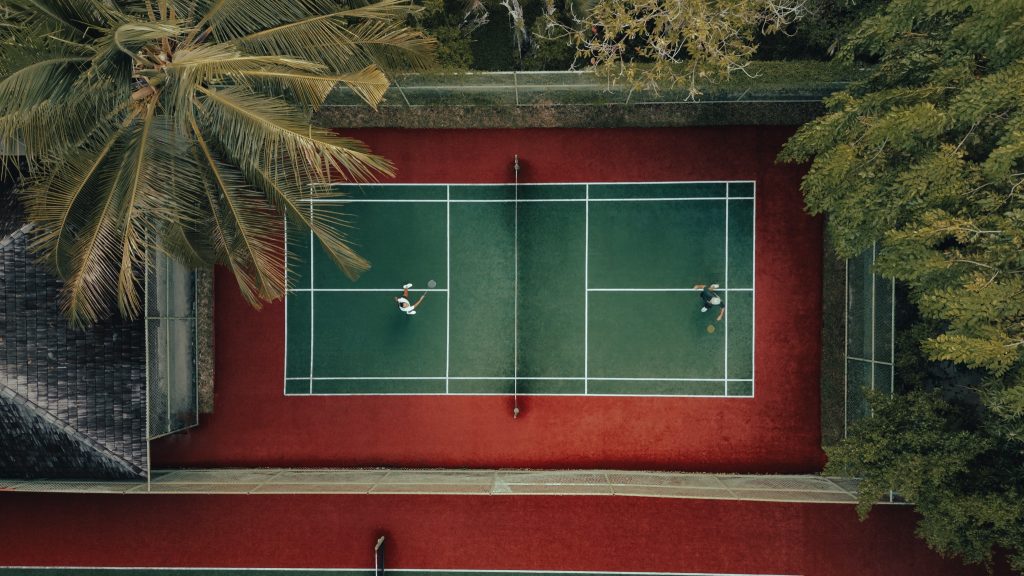 Tennisplatz mit grünem synthetischem Belag und roten Linien, umgeben von Bäumen und Palmen auf den