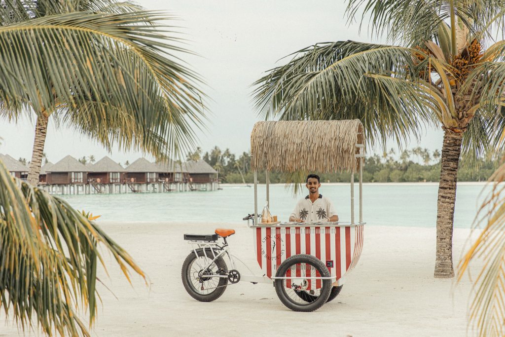 Strand mit weiß-rotem Fahrradstand mit Strohdach, Palme und Bungalows auf Stelzen im Wasser auf den