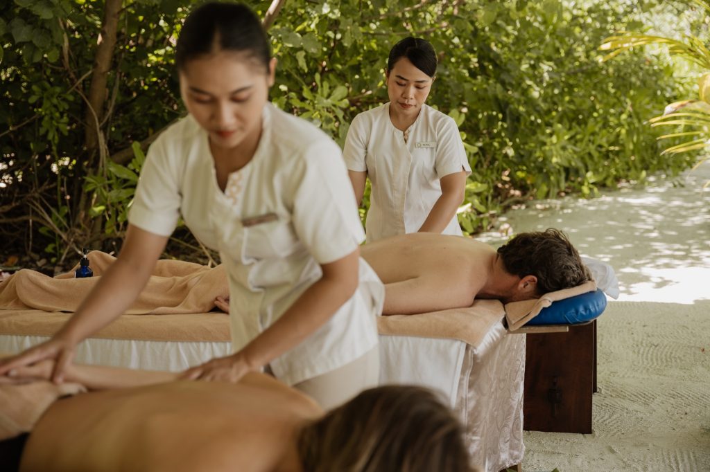 Zwei Personen liegen auf Massagebetten im Freien, umgeben von tropischem Grün, Masseure in weißen Uniformen