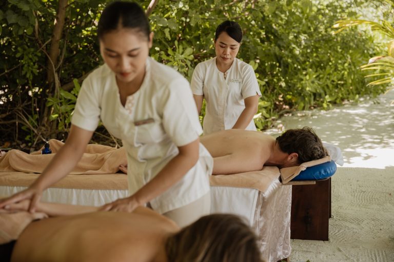 Zwei Personen liegen auf Massagebetten im Freien, umgeben von tropischem Grün, Masseure in weißen Uniformen