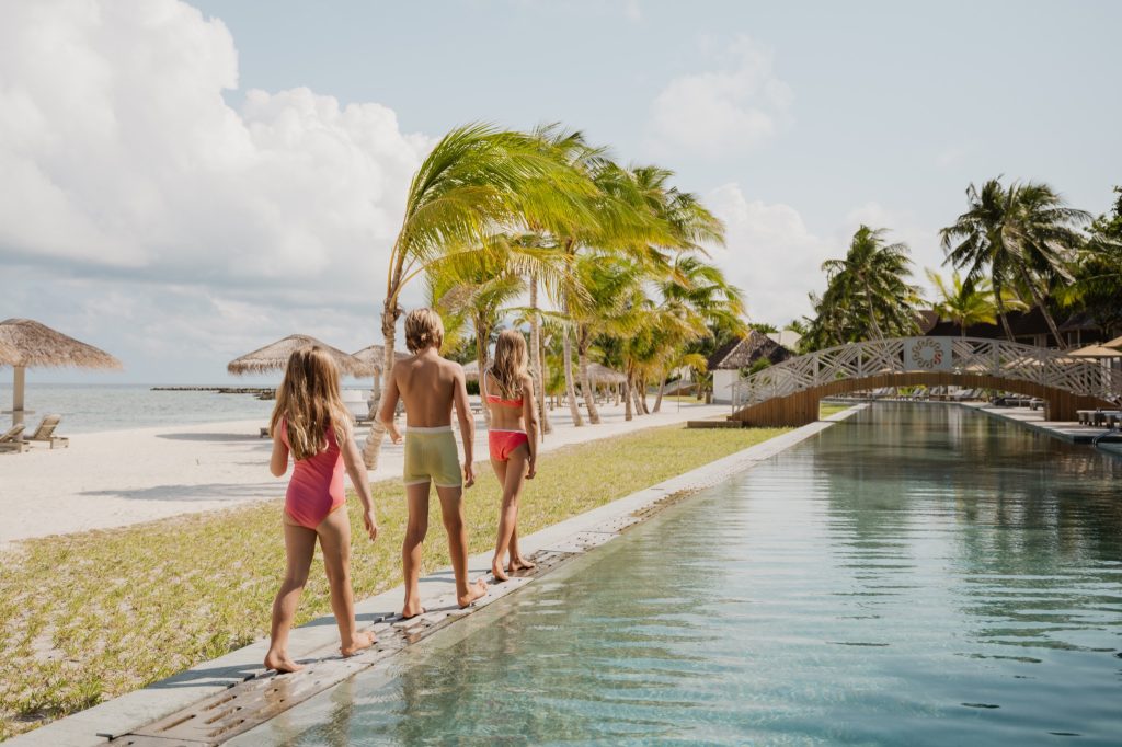 Poolbereich mit Palmen, mehreren Personen am Pool, weißer Sandstrand, Holzhütten und Liegestühle im Hintergrund auf den.