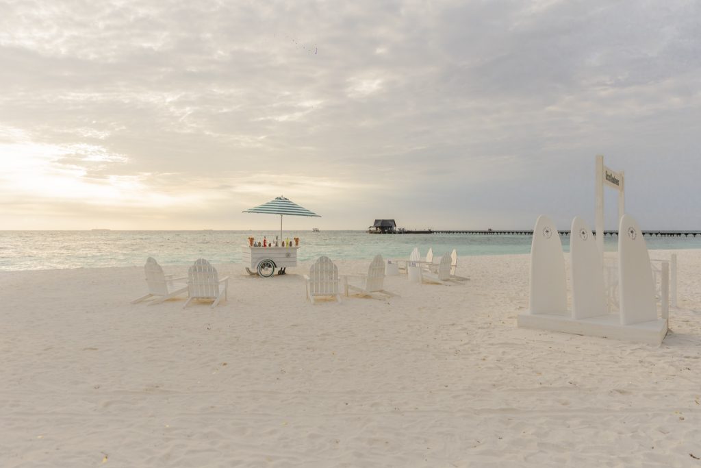 Strand mit hellem Sand, weißen Liegestühlen, gestreiftem Sonnenschirm und türkisblauem Ozean im Hintergrund auf den
