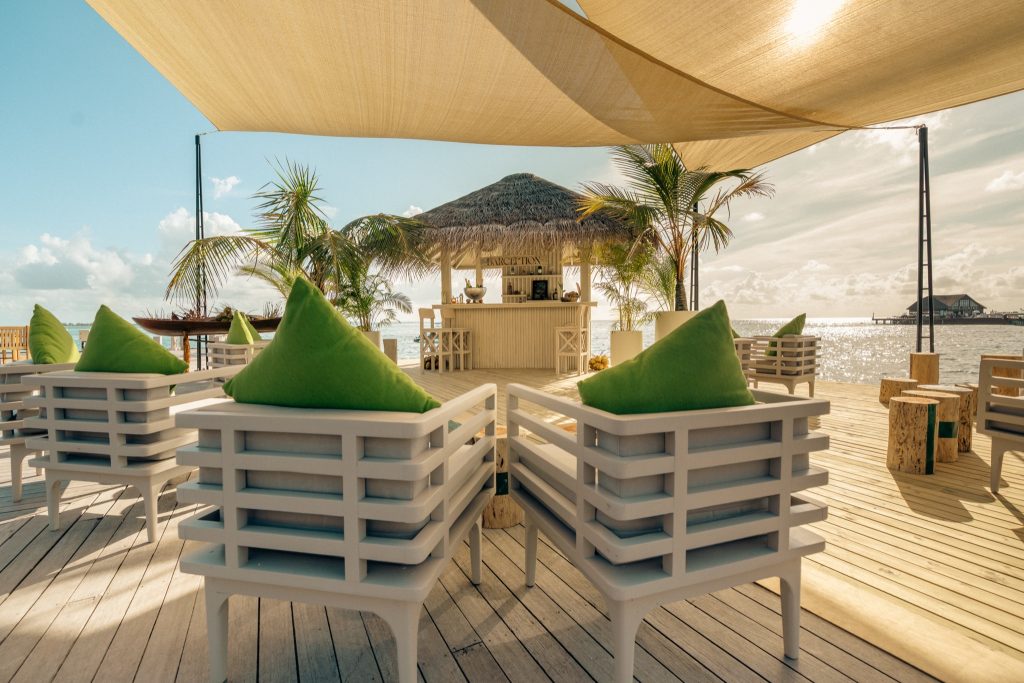 Strandbar Barception mit strohgedecktem Dach, weißen Holzstühlen und grünen Kissen, Palmen und Ozean im Hintergrund
