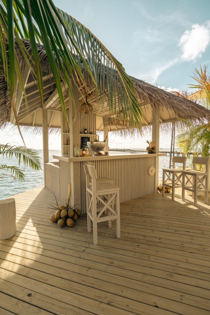 Strandbar aus hellem Holz mit Strohdach, Holzstühlen und Meerblick bei Sonnenschein auf den