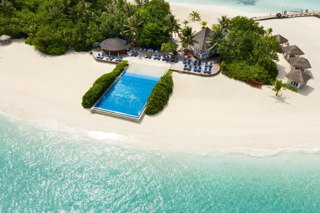 Vogelperspektive auf Sun Siyam Olhuveli Infinity-Pool mit weißem Sandstrand und blauem Meer, umgeben von Vegetation