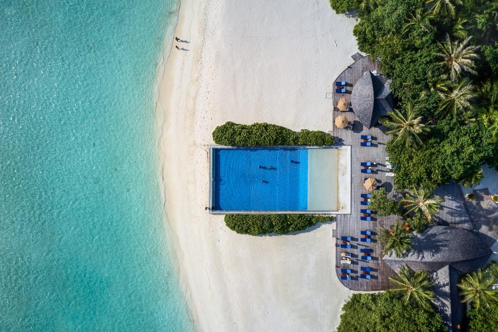 Luftaufnahme des rechteckigen Swimmingpools mit Holzdeck und Liegestühlen im Sun Siyam Olhuveli auf den
