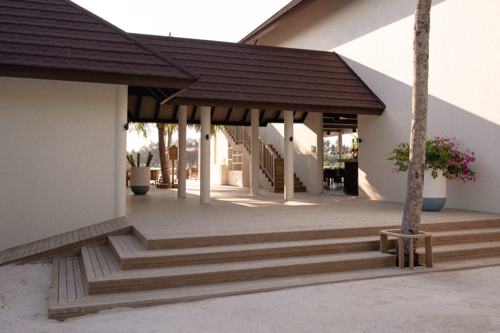 Offener Außenbereich der Bar Olive im Sun Siyam Olhuveli mit Holztreppe, hellen Gebäuden und großen Pflanzentöpfen