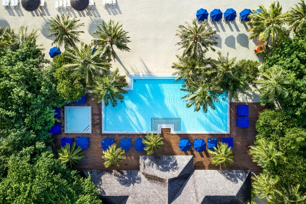 Vogelperspektive auf den rechteckigen Pool mit blauem Wasser, Holzdeck, Palmen und weißem Sandstrand im Hintergrund