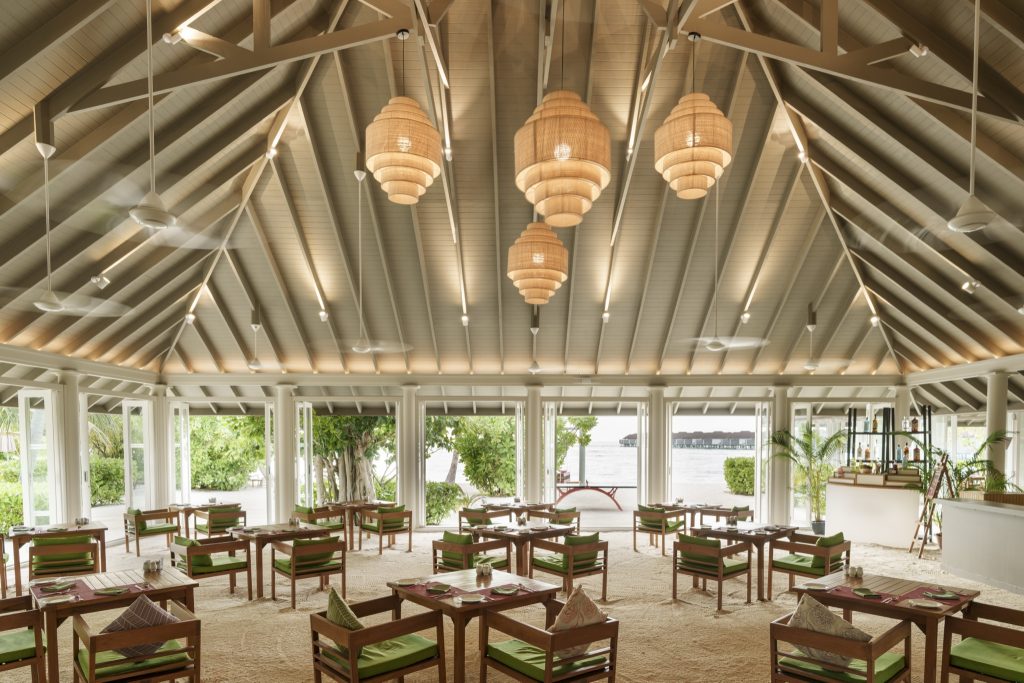 Helles Restaurant Malaafaiy mit Holzdecke, großen Fenstern, grünen Polstern und Blick auf Garten und Meer