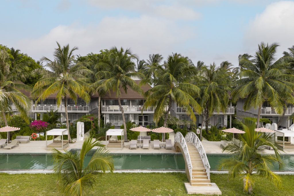 Langgezogener Pool mit Holzbrücke, Liegestühlen unter beige-weißen Sonnenschirmen und Palmen im Sun Siyam Olhuveli