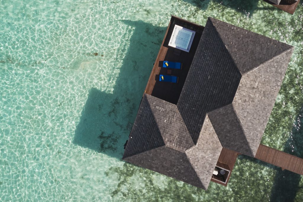 Vogelperspektive auf Sun Siyam Olhuveli Prestige Jacuzzi Water Villa mit Holzterrasse, zwei blauen Liegestühlen und Jacuzzi.