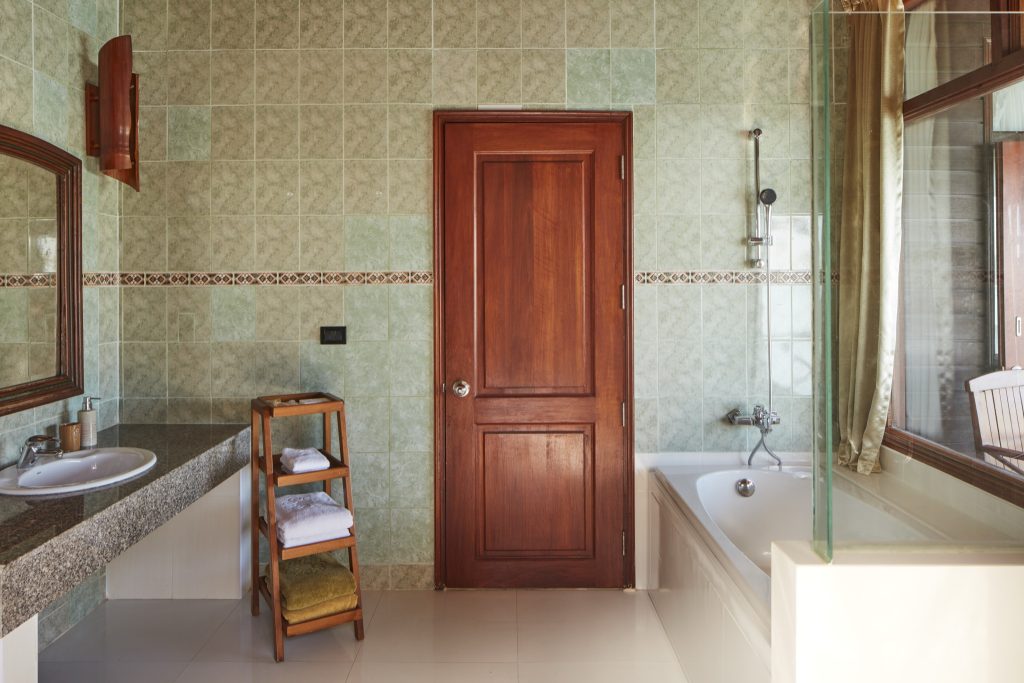 Badezimmer der Water Villa im Sun Siyam Olhuveli mit grünen Wandfliesen, Holztür, Waschtisch und Badewanne mit Glastrennwand