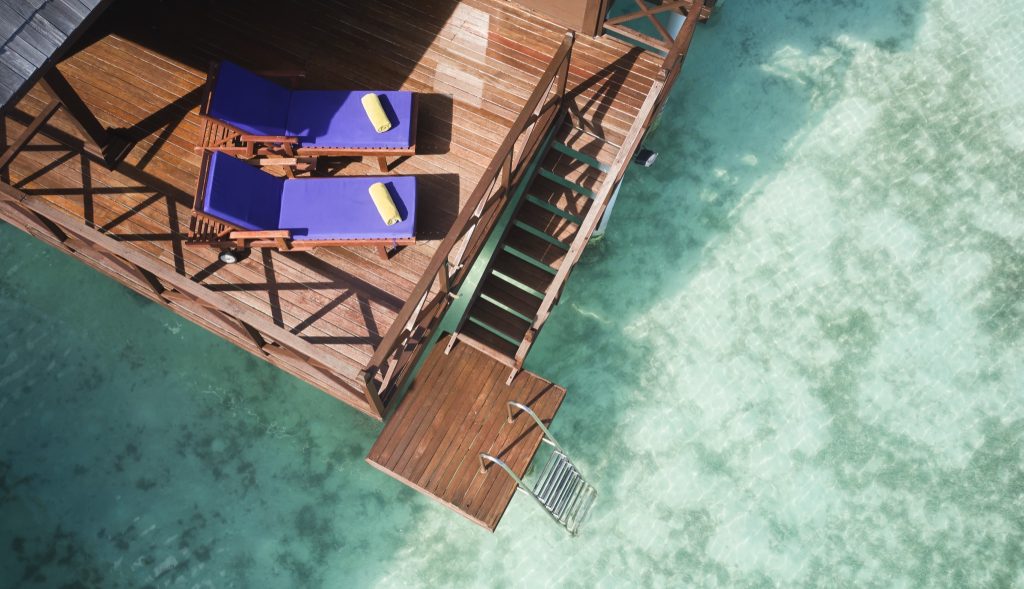 Holzplattform der Water Villa mit zwei blauen Liegestühlen und gelben Handtüchern über türkisfarbenem Wasser
