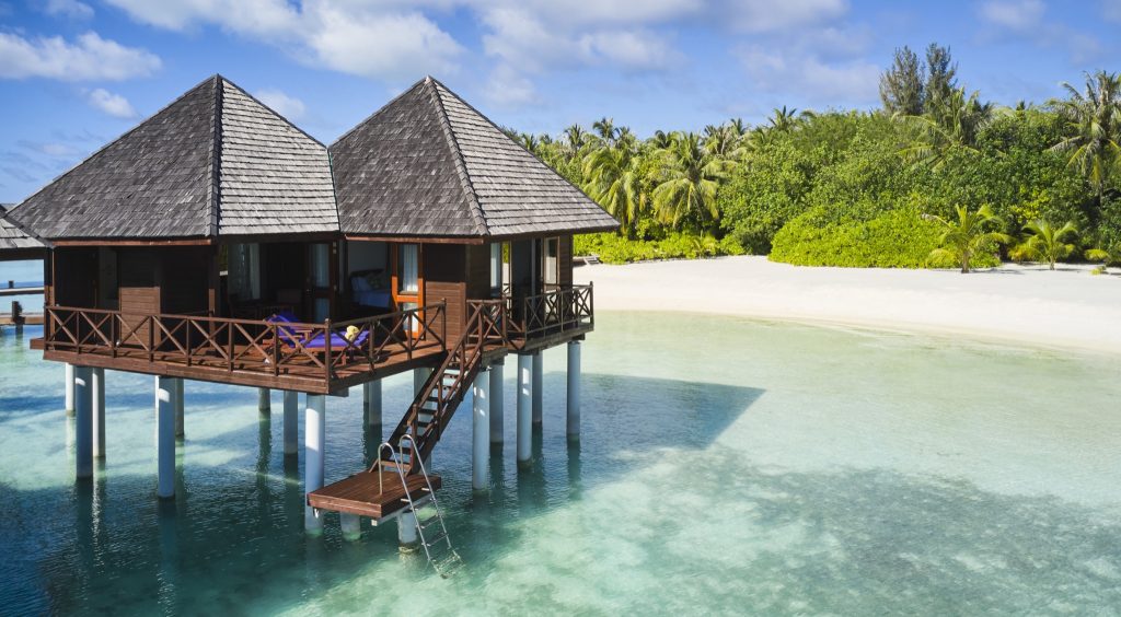 Hotelbungalow auf Stelzen über türkisfarbenem Wasser mit Holztreppe und Metallgeländer, Palmen im Hintergrund
