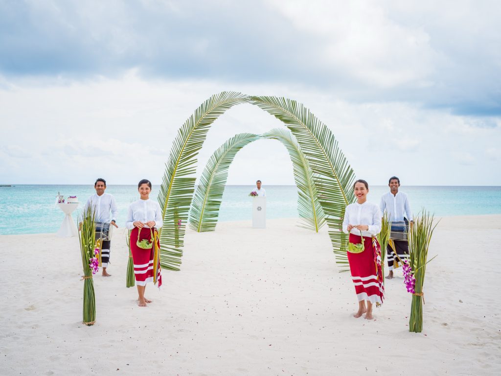Hochzeitskulisse am Strand bei Vakkaru Maldives mit Palmenbogen, festlich gekleideten Personen und türkisfarbenem Meer