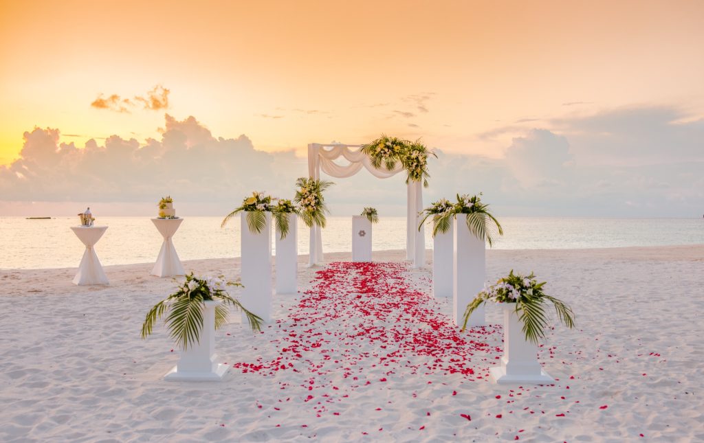 Strandhochzeit mit weißen Säulen, Traubogen, roten Blütenblättern und orangefarbenem Himmel auf den