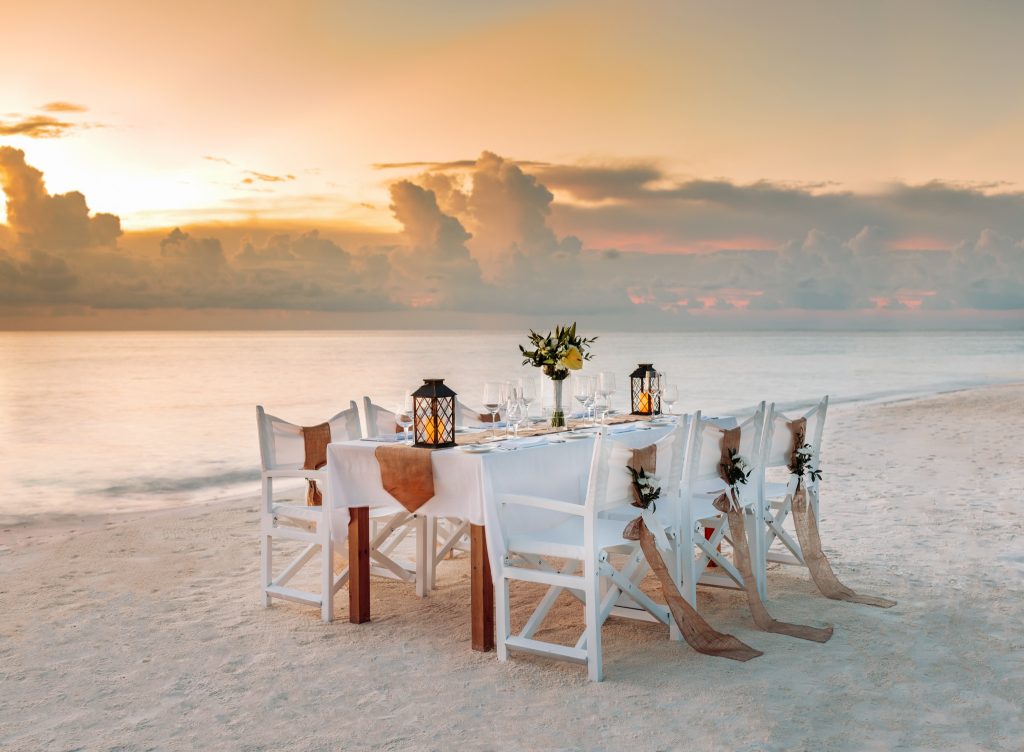 Weiß gedeckter Esstisch mit Holzmöbeln, Laternen und Blumen am Strand mit Meer im Hintergrund