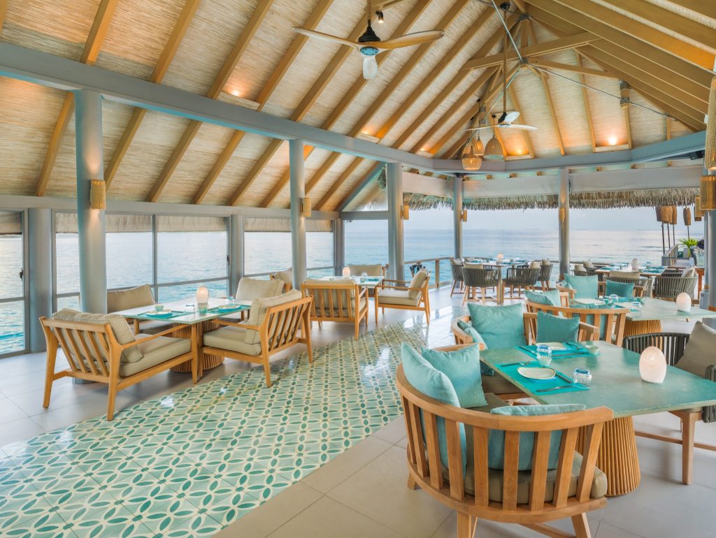 Offener Barbereich Vakkaru Maldives Bar Lagoon mit Holztischen, türkisfarbenen Kissen und Blick auf das Meer