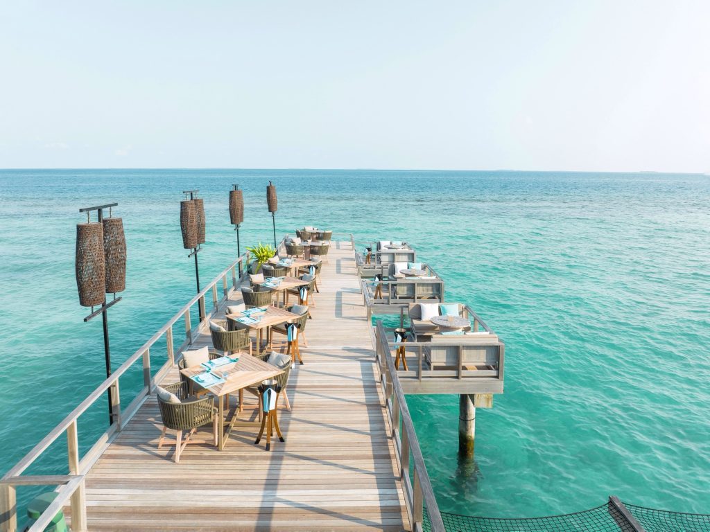 Holzterrasse mit Tischen, Stühlen und hohen Lampen über türkis-blauem Meer bei hellem Tageslicht