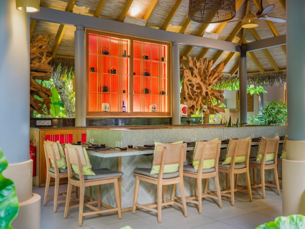 Barbereich im Vakkaru Maldives mit grauer Stein-Theke, hellen Holzstühlen und orangefarbener Holzregalwand