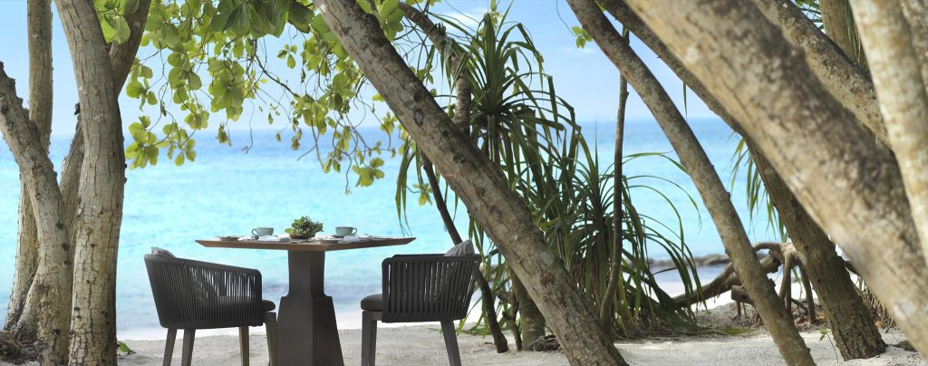 Essbereich am Strand mit Holztisch, grauen Sesseln, Kaffeetassen und tropischen Bäumen im Hintergrund