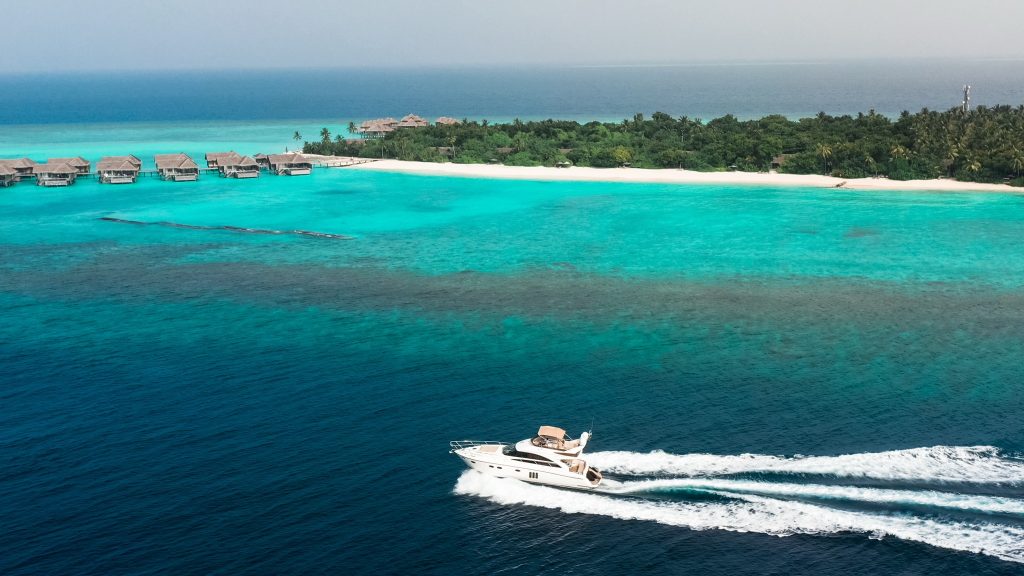 Wasserbungalows auf Pfählen im türkisblauen Meer mit weißer Yacht und grüner Insel im Hintergrund