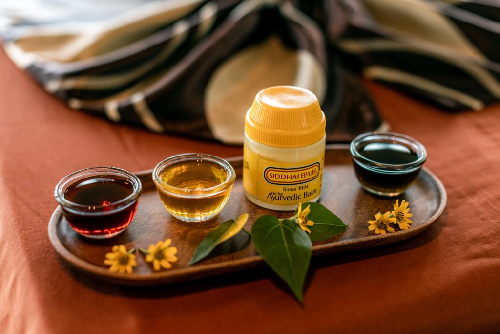 Drei kleine Glasschalen mit farbigen Flüssigkeiten und ein gelber Ayurvedic Balm auf Holztablett mit Blättern